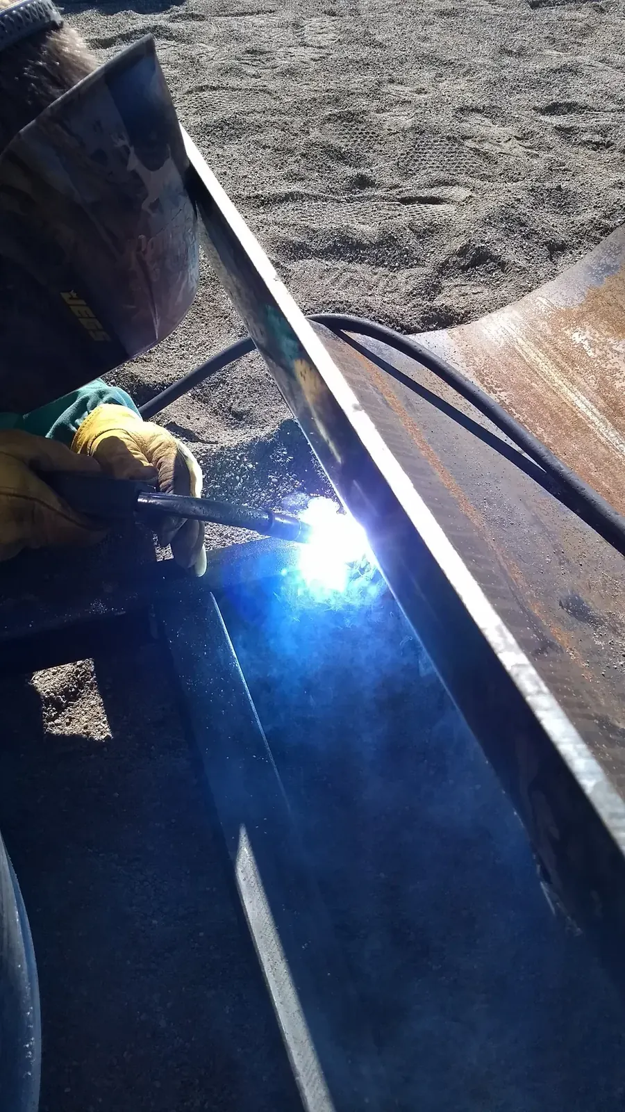 Close view of a welder joining a curved steel pipe section to its support during outdoor installation.