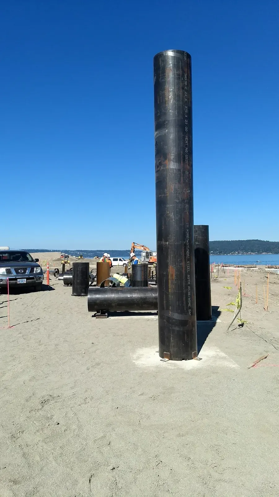 The intact vertical pipe shown from its full top to its base, with shorter steel sections and workers behind.