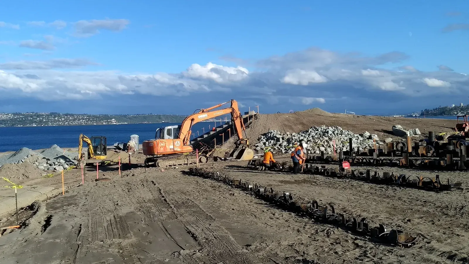 Workers among long rows of small steel pipe fragments at the waterfront construction site.