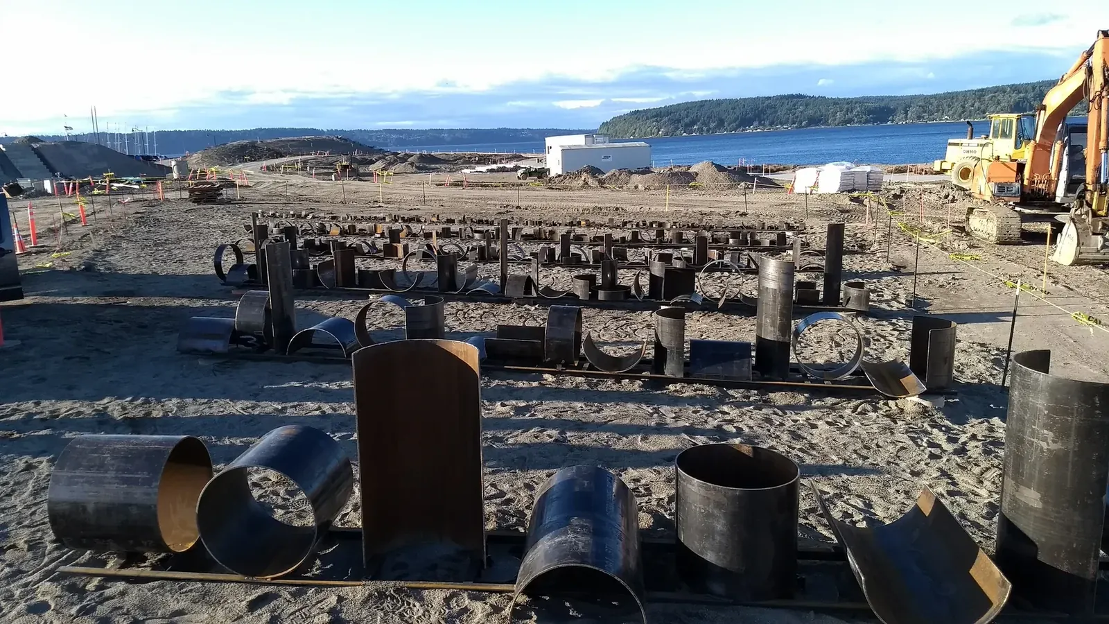Circular and curved steel pipe sections in the foreground, with progressively smaller rows extending toward the water.