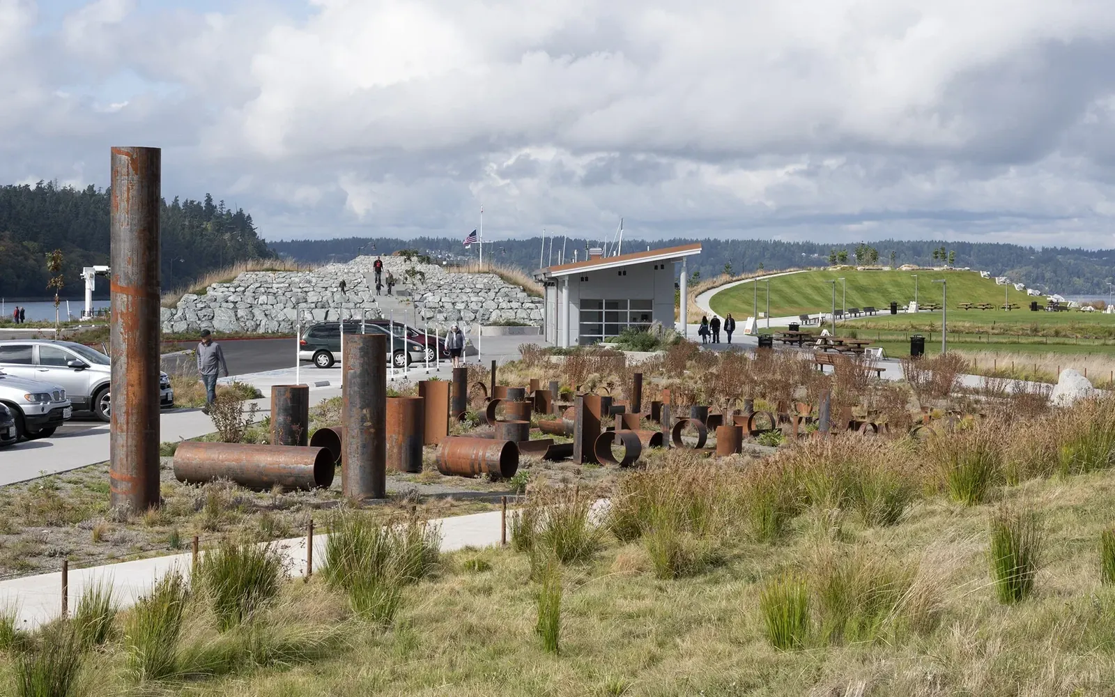 Alluvion in the landscaped park, with the full upright steel pipe at left and rows of smaller weathered pieces among paths and grasses.