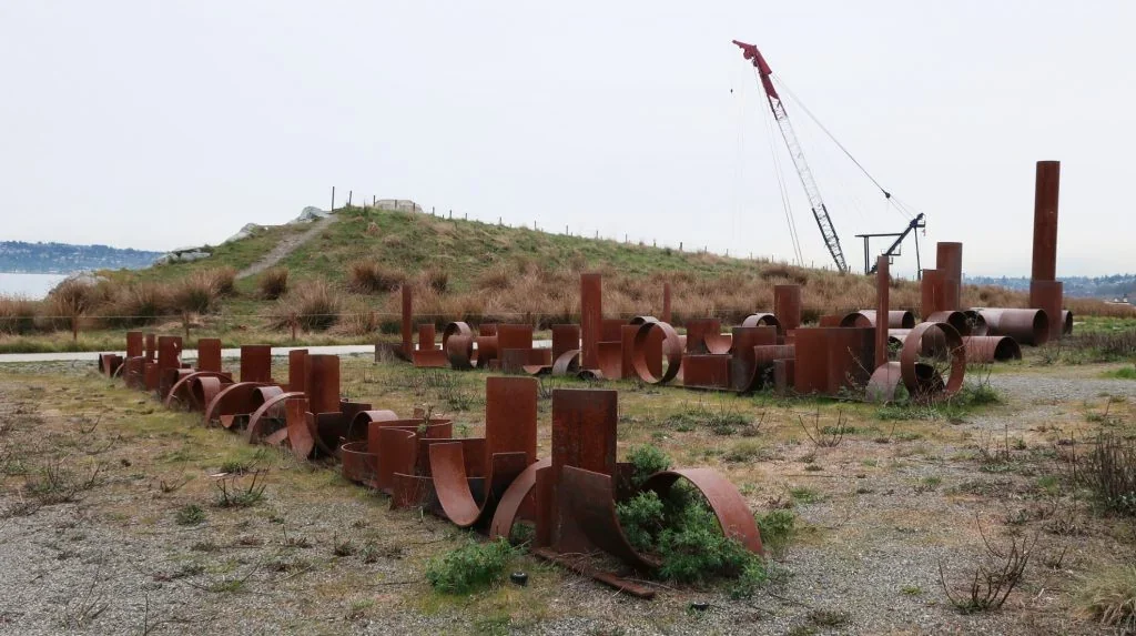 Low curved and angular steel pieces across the foreground, with taller pipes beyond, park paths, a grassy mound and a crane in the distance.