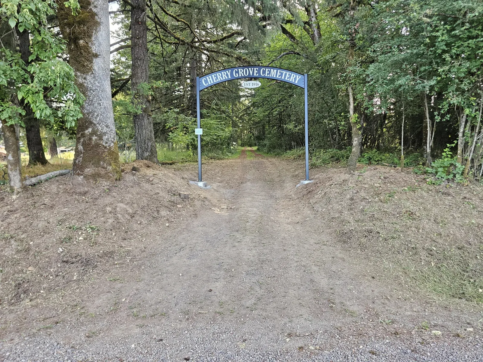 A wide view of the cleared cemetery driveway, flanked by trees and marked by the new arched sign.