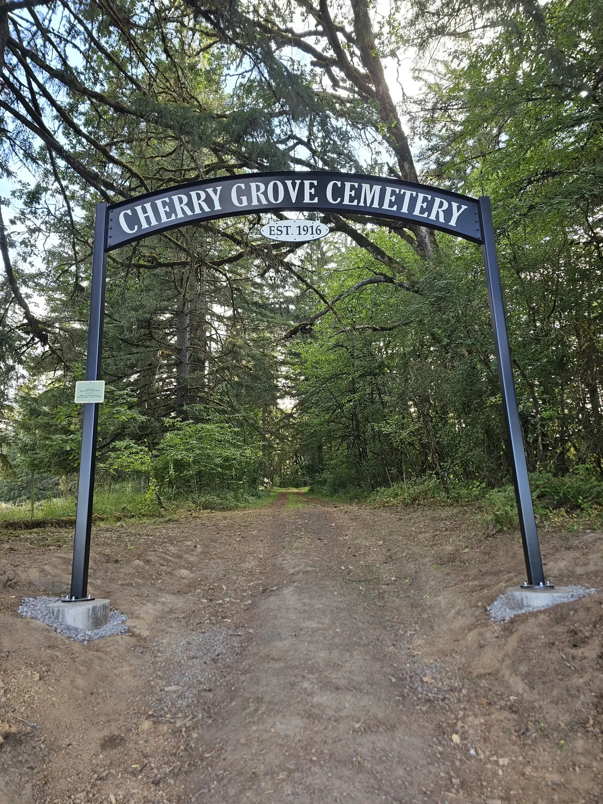 The completed arched Cherry Grove Cemetery sign and two posts frame the cleared driveway through the trees.