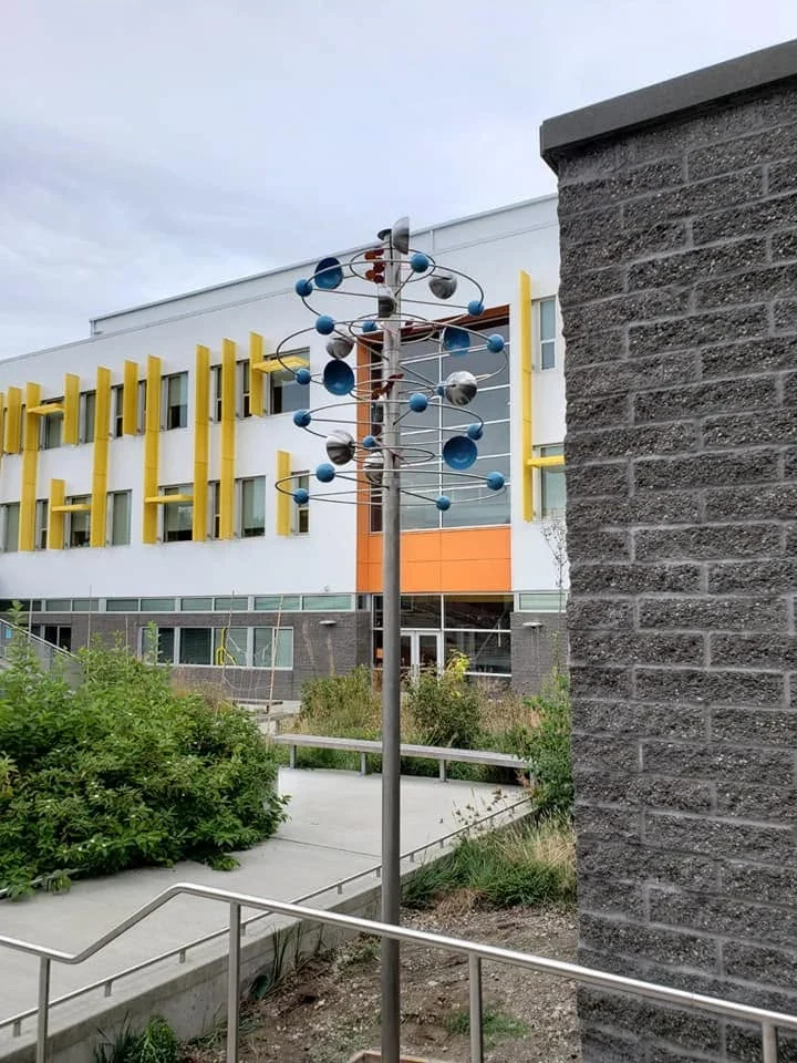 Carbon Atom Whirligig beside the school courtyard stairs, with the school building behind it.