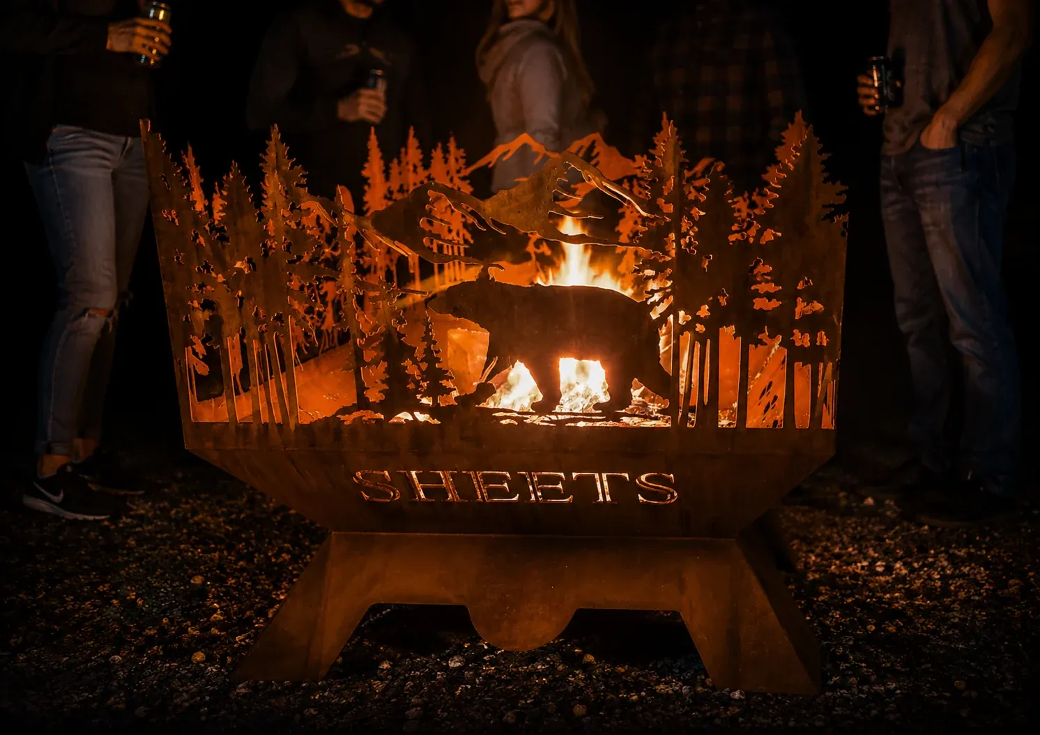 A steel fire pit burning outdoors, with bear silhouettes and Sheets lettering cut into the side.