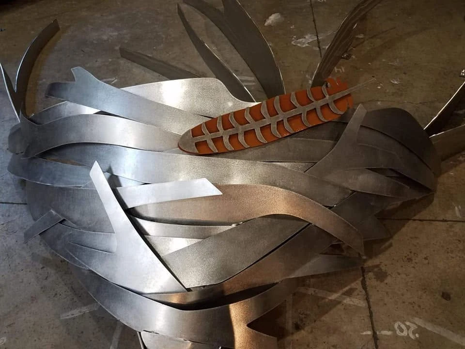 Overlapping curved stainless steel strips around the orange center of Fledge, photographed on the shop floor.