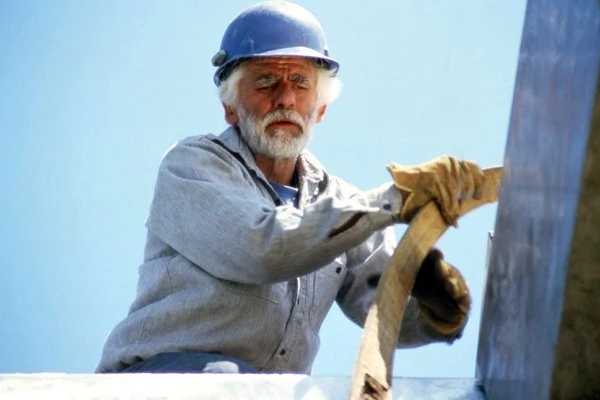 Lee Kelly in a blue hard hat and work gloves handling a lifting strap beside a steel sculpture.