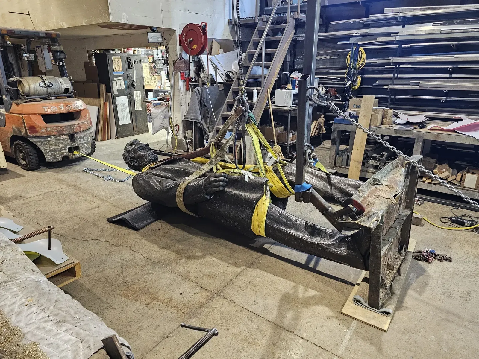 Lincoln lying horizontally in the shop, supported by lifting straps, chains, and a fabricated frame around the bent base.