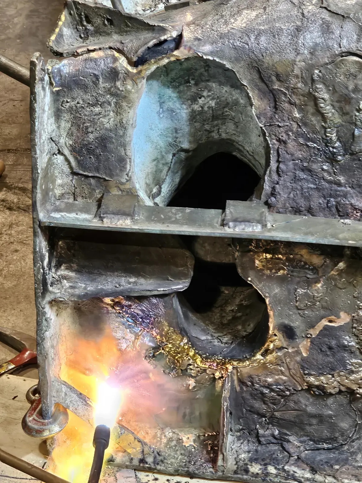A torch heating the underside of the original bronze base, with cracks and earlier welds visible around the foot openings.