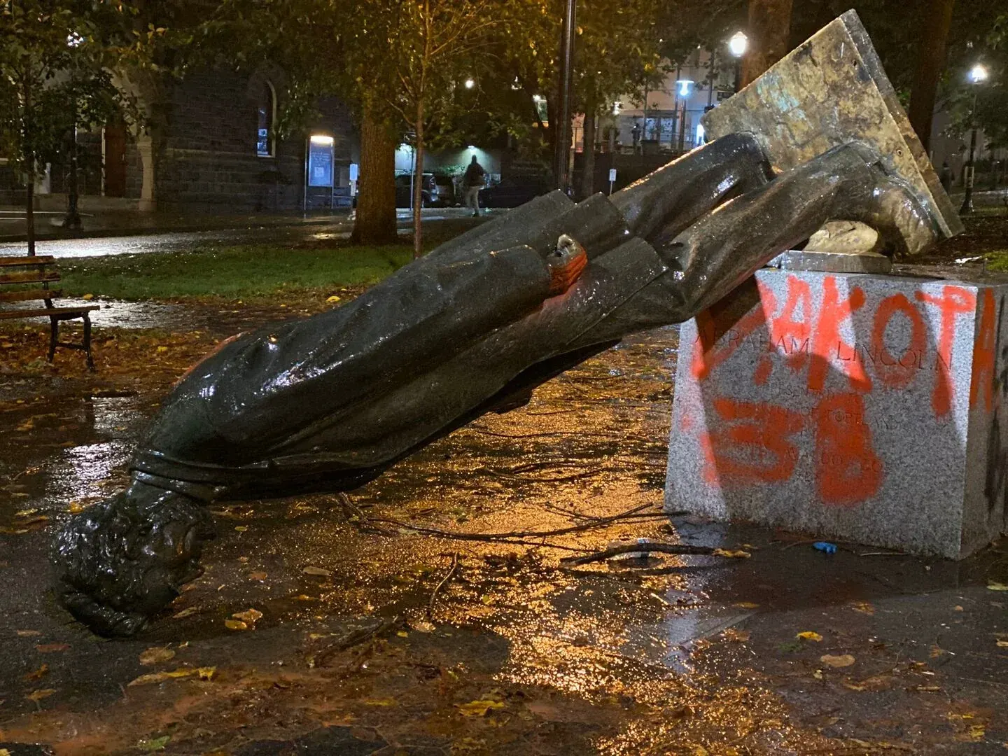 Abraham Lincoln statue lying head down beside its pedestal at night, with its damaged base raised in the air.