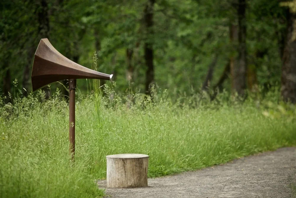 A rusted steel Listening Trumpet with a small stainless steel earpiece beside a gravel trail and tall green grass.
