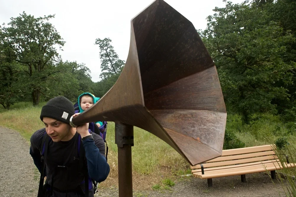 A visitor carrying a child listens at the narrow stainless steel end of a broad, rusted steel trumpet.