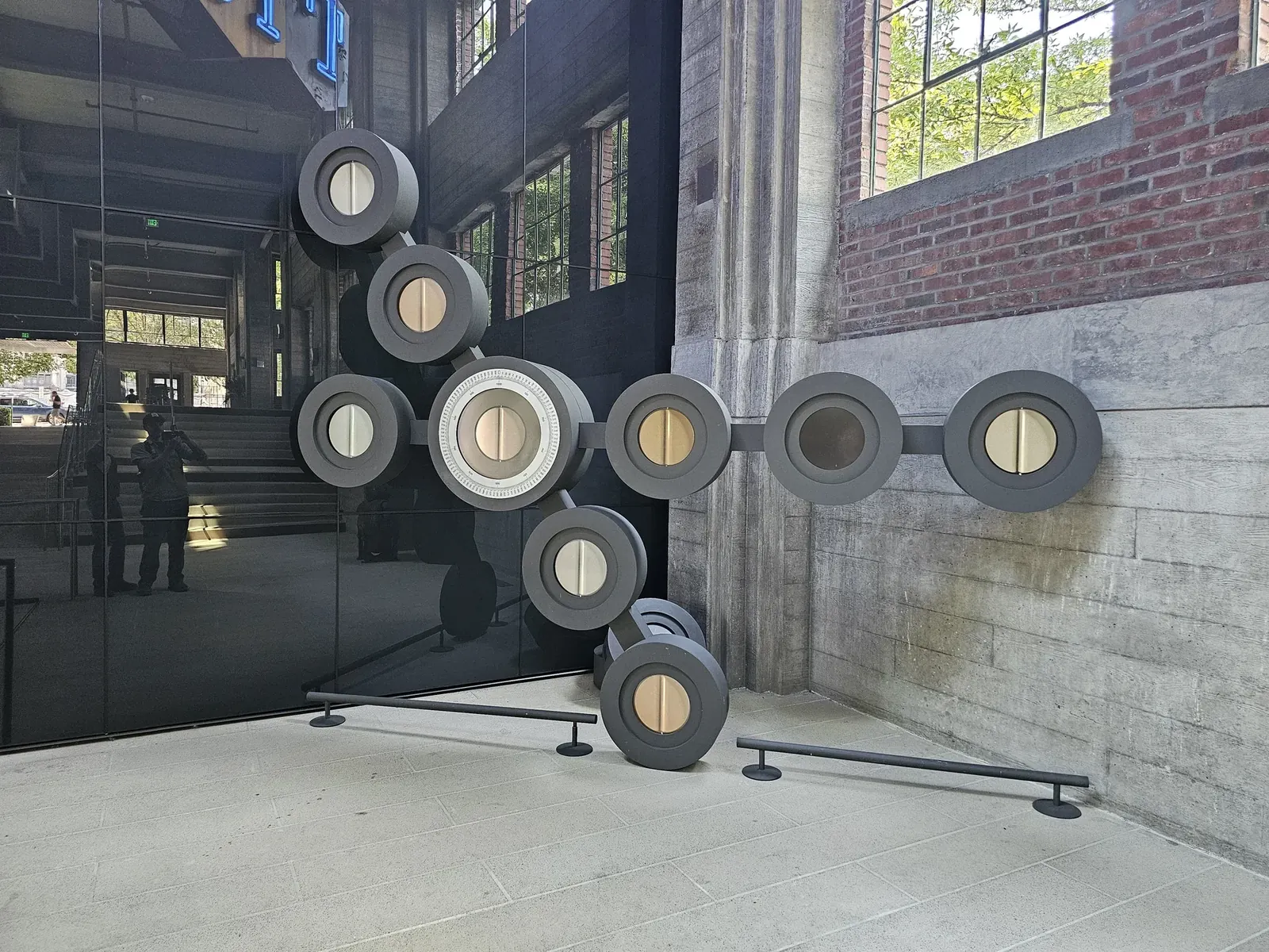 The complete Mechanism sculpture with circular display housings on crossing aluminum arms, standing beside the historic brick and concrete walls at Troy Block.
