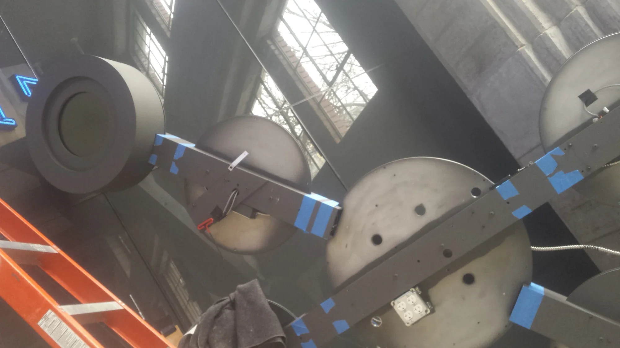 Close-up from below of the circular aluminum housings, rectangular connecting arms, openings and exposed wiring during assembly.