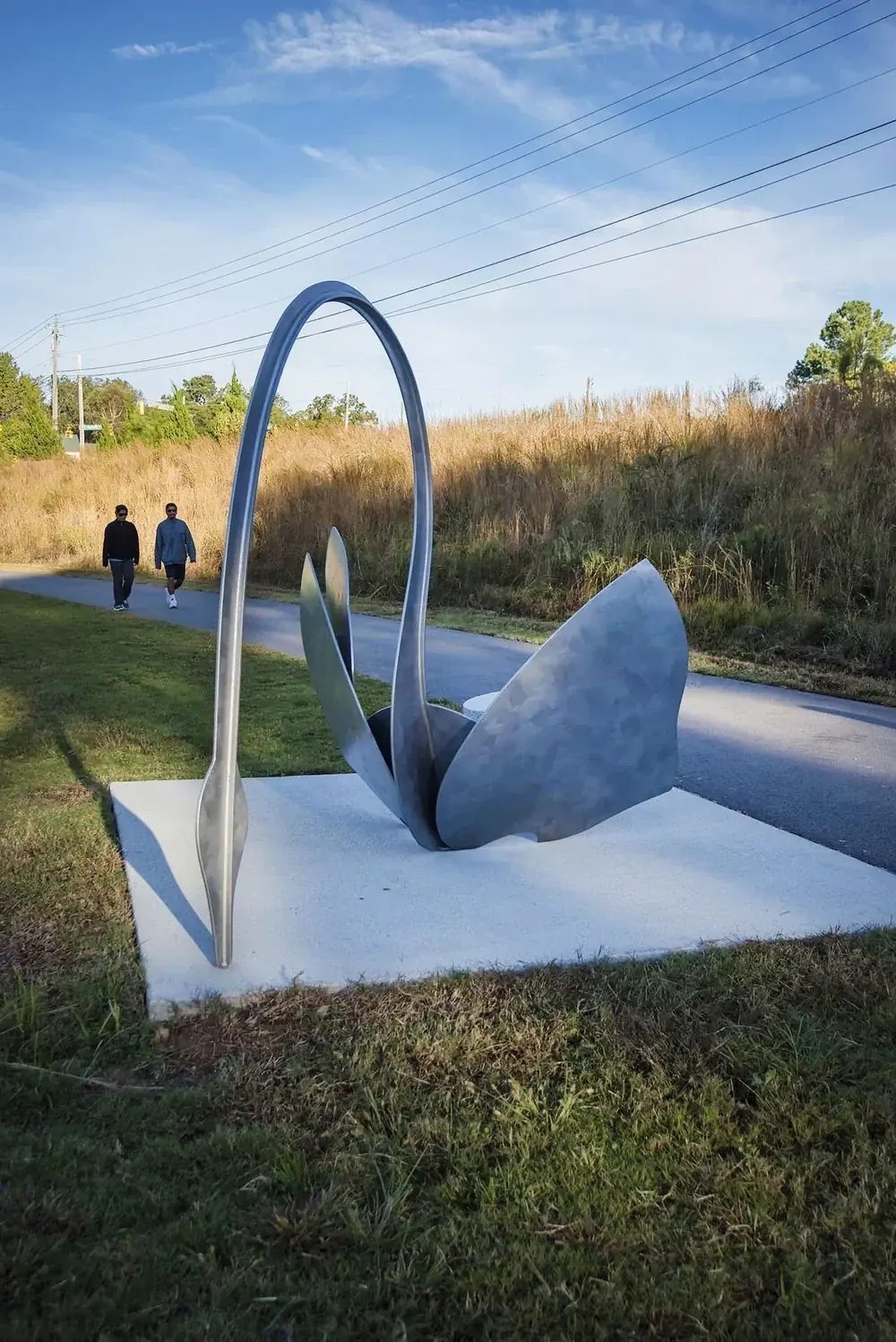Side view of a steel swan showing the full arch of its neck and two broad plate wings, with visitors walking on the path behind it.