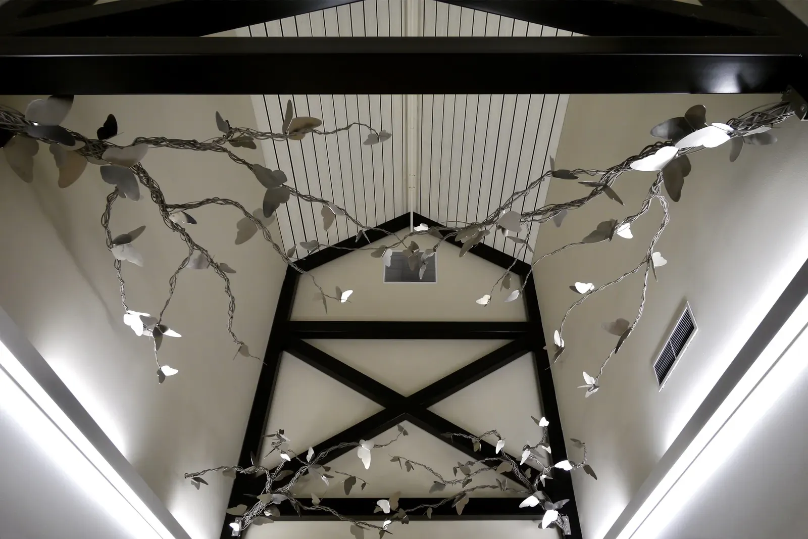 Wide view of reflective stainless steel butterflies along twisting branches suspended between the dark beams of a vaulted school ceiling.