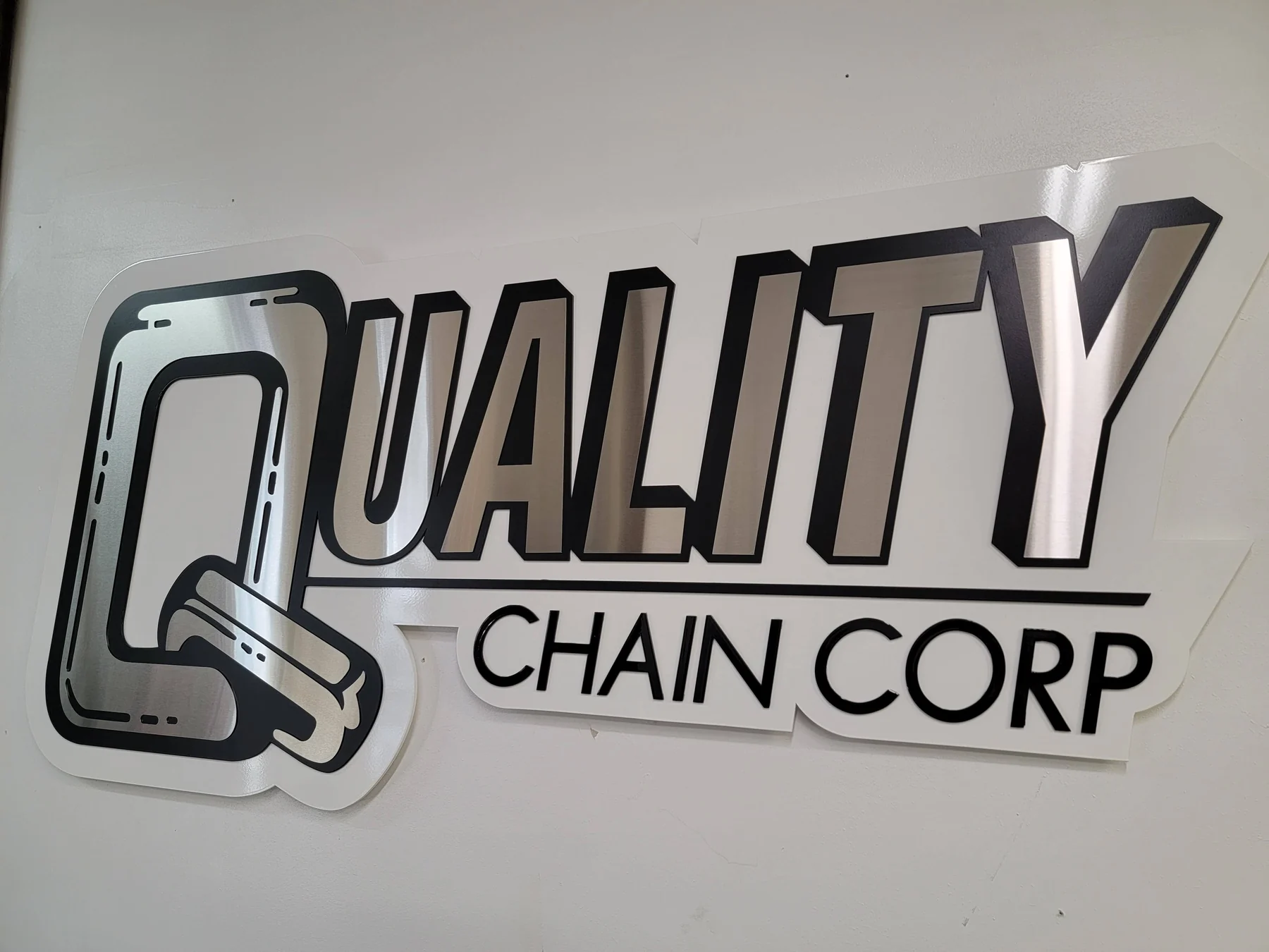 Layered Quality Chain Corp metal logo and lettering on a light wall