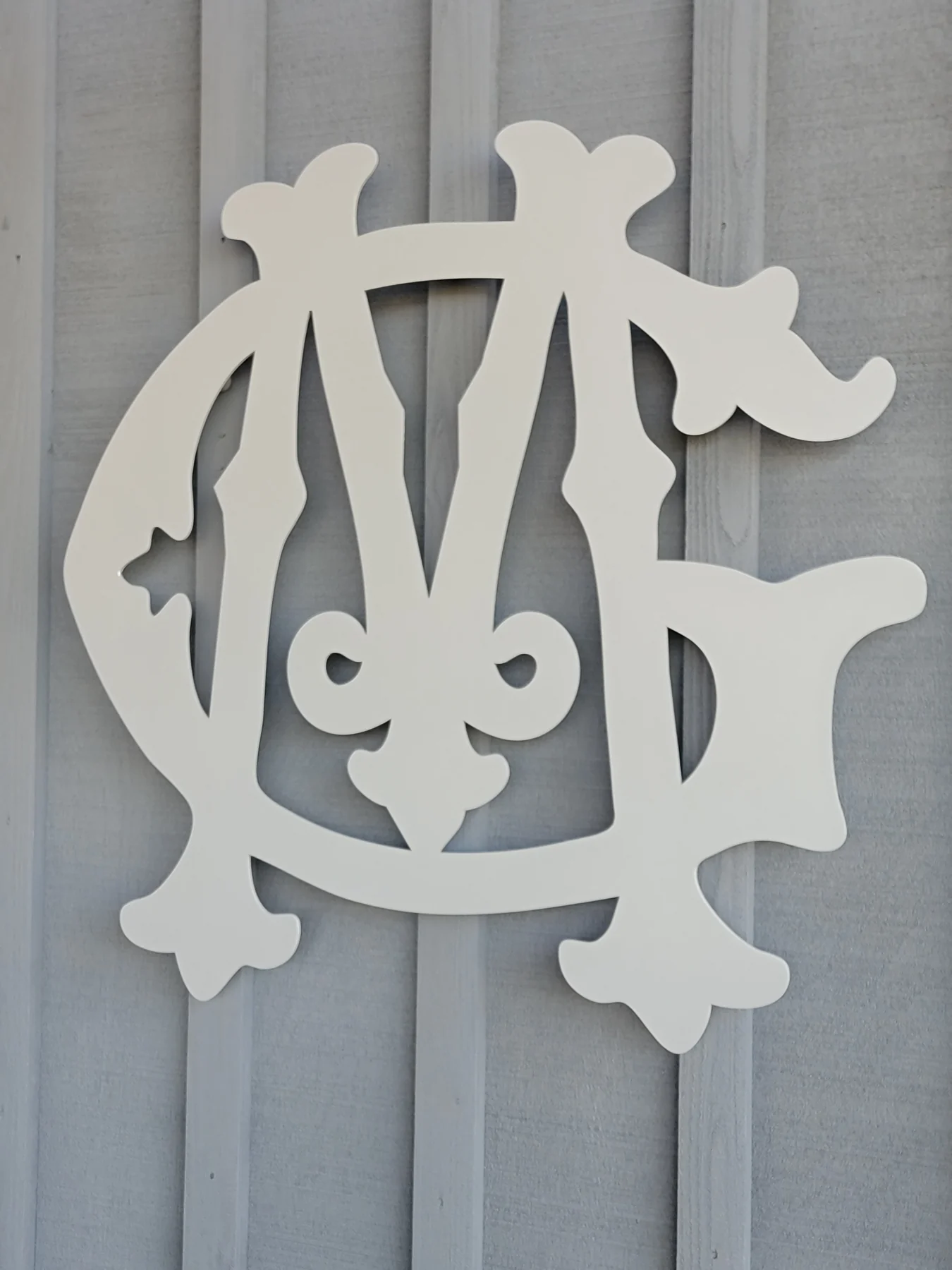 Light-colored ornate interlocking metal letters on gray exterior siding
