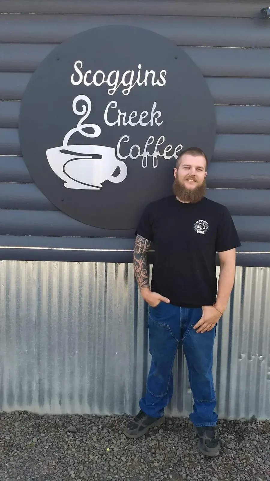 Large round Scoggins Creek Coffee sign with light script and a coffee-cup graphic, with a person standing beside it