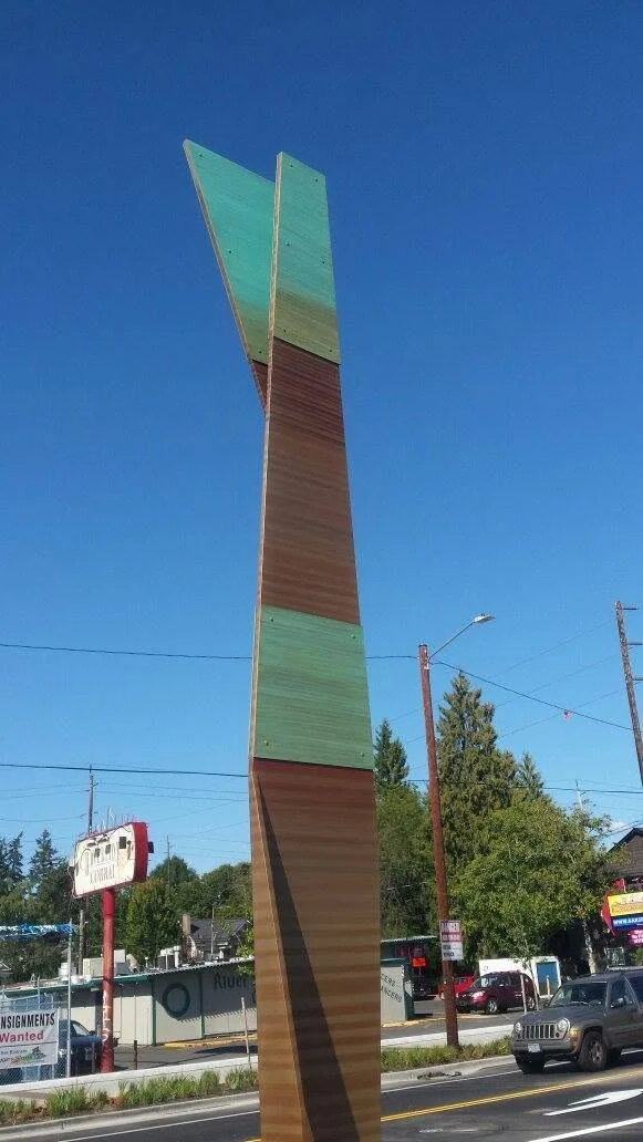 Tall folded metal totem with layered bronze-colored surfaces and turquoise accents beside Sellwood Bridge.