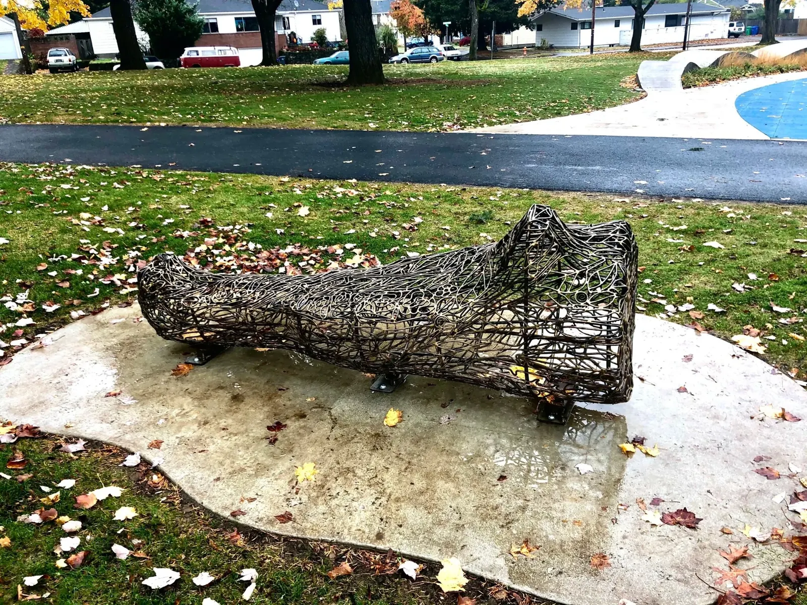 A reclining bronze log form, made with open rod patterns, on a concrete pad beside the playground.