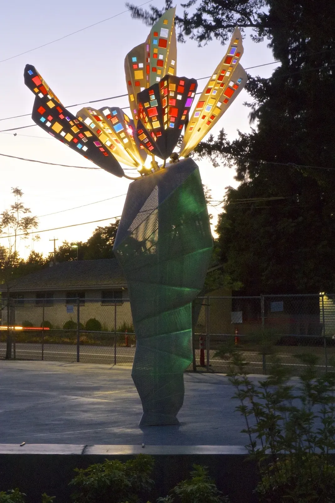 The Fifth Wind illuminated at dusk, with five colored-glass wings above the perforated stainless steel base.