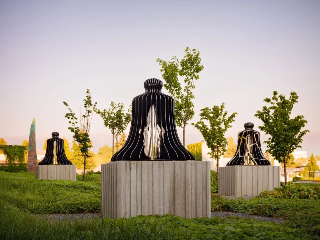 Three black bell sculptures on concrete plinths among trees and planting at Alder Passage, with face profiles visible in the nearest bell.