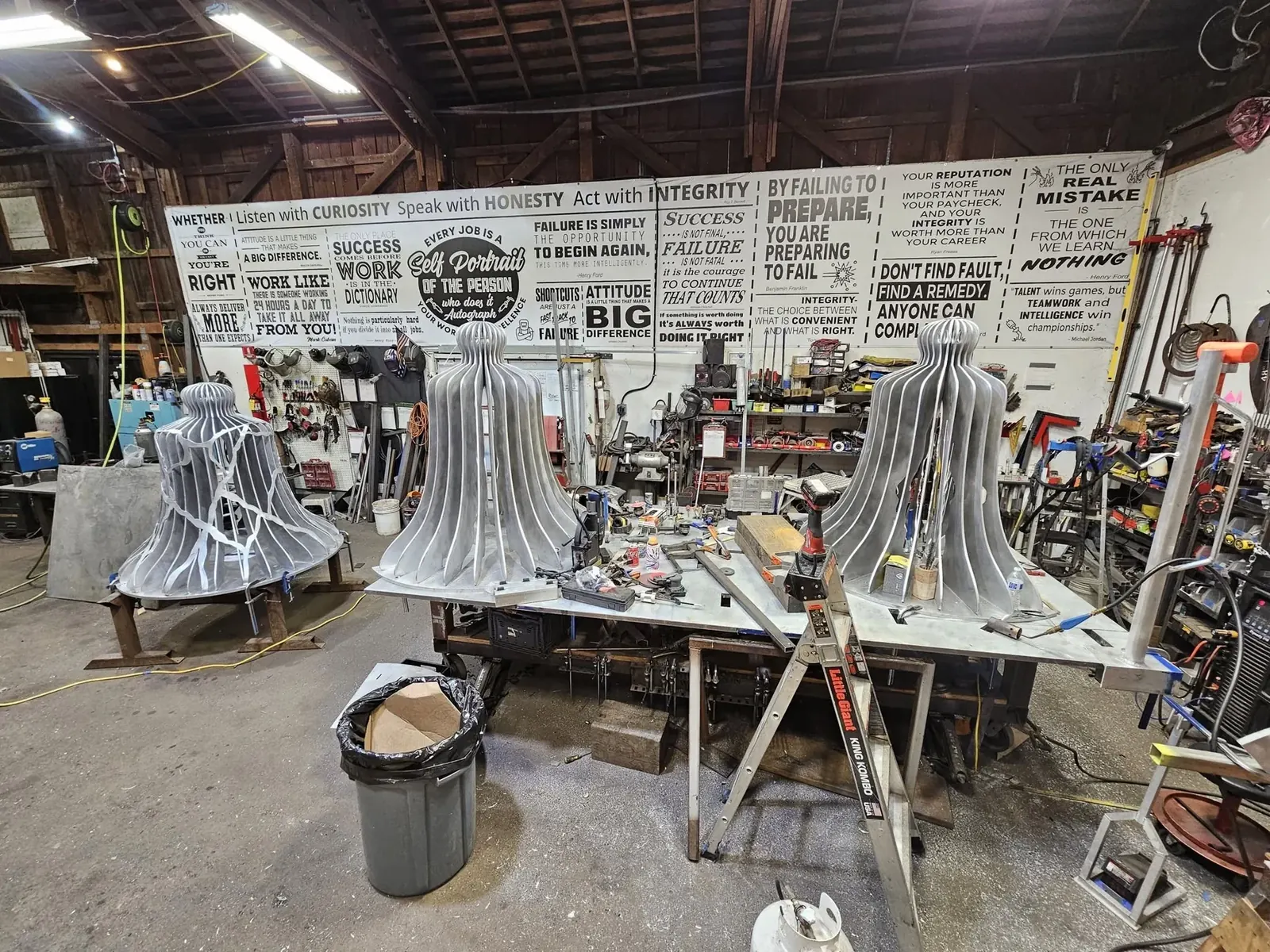 Three unpainted aluminum bell sculptures standing on fabrication tables inside Western Iron Works.