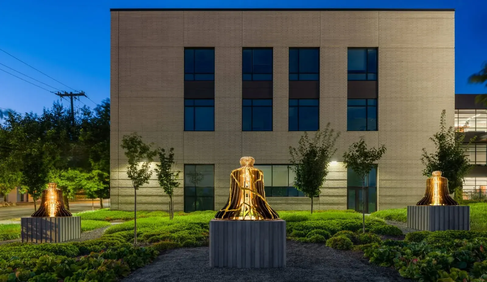 Three illuminated bell sculptures on concrete bases along a landscaped path outside the justice center at dusk.