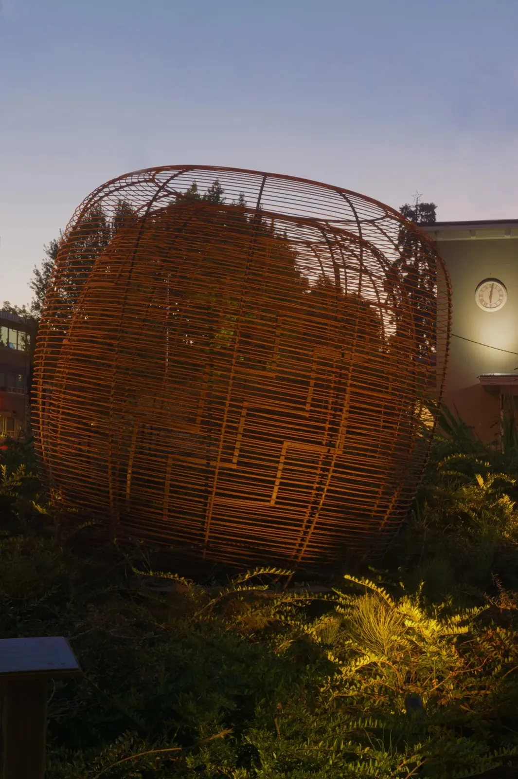 The broad basket sculpture at dusk, its steel lines and inner bird motif illuminated against the evening sky.