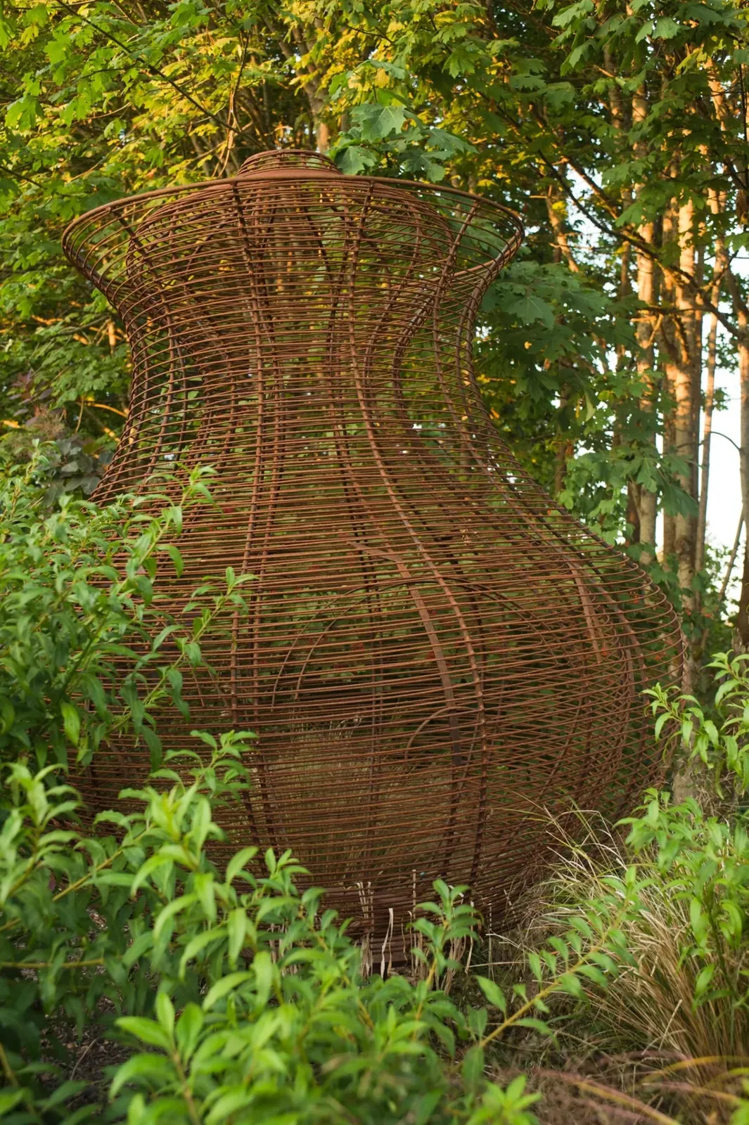 Steel basket sculpture with a flared upper rim and narrow waist, set among green plants at Waypoint Park.