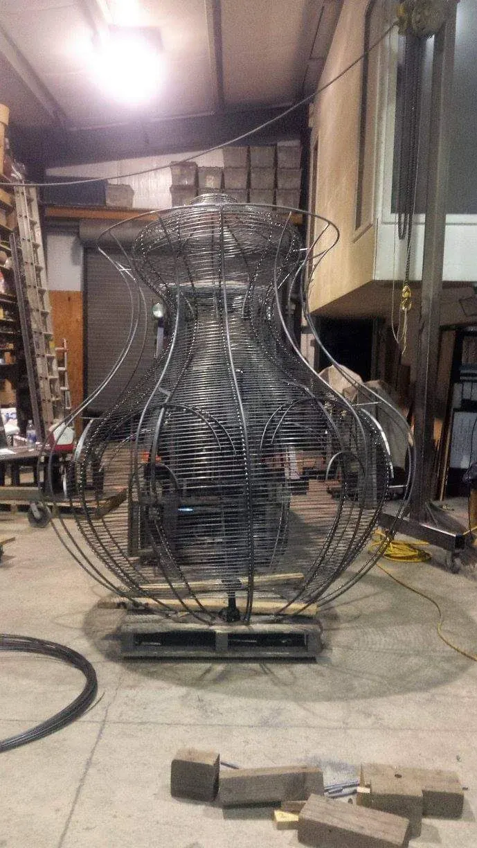 A flared basket form on a wheeled platform in the shop, with curved vertical ribs and closely spaced horizontal steelwork.
