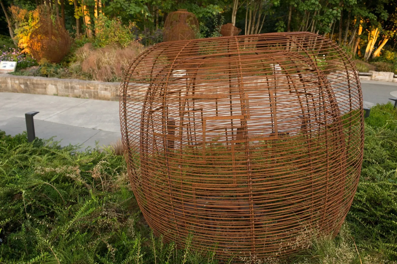 Wide, low steel basket sculpture beside the park path, with an inner form visible through its horizontal steelwork.