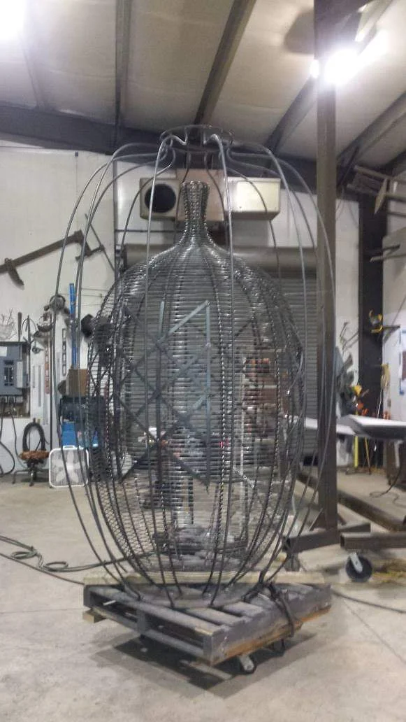 Tall basket framework on a wheeled support in the shop, with a smaller patterned vessel visible inside.