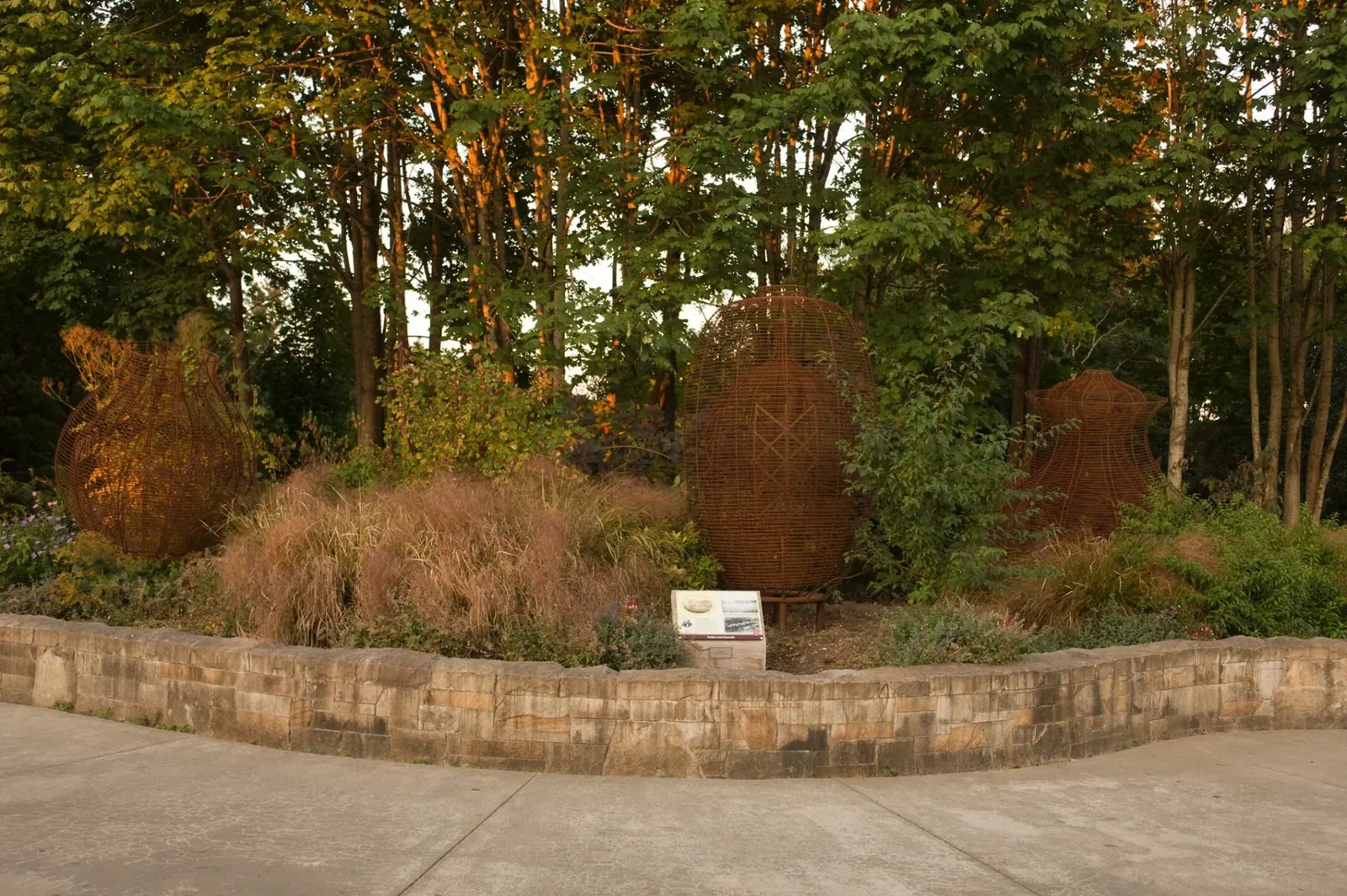 Three of the four rust-colored steel basket sculptures among the trees and planting beds at Waypoint Park.