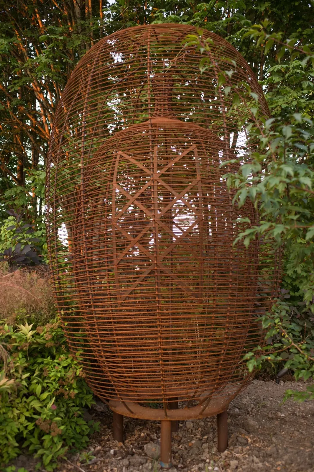 Tall steel basket sculpture with a smaller inner vessel and an angular rune pattern, surrounded by foliage at Waypoint Park.